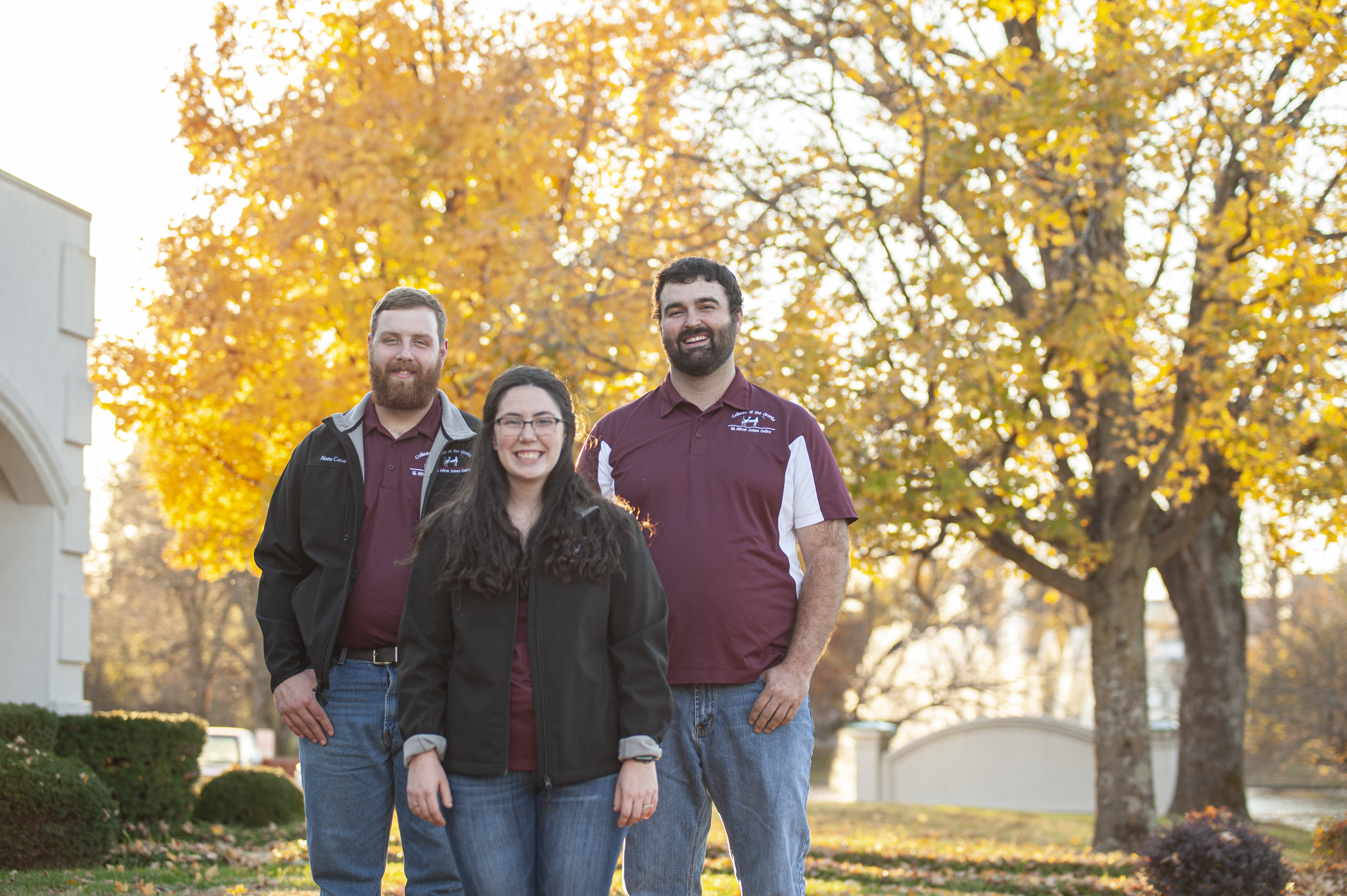 A woman stands in front of two men by a yellow-leafed trees